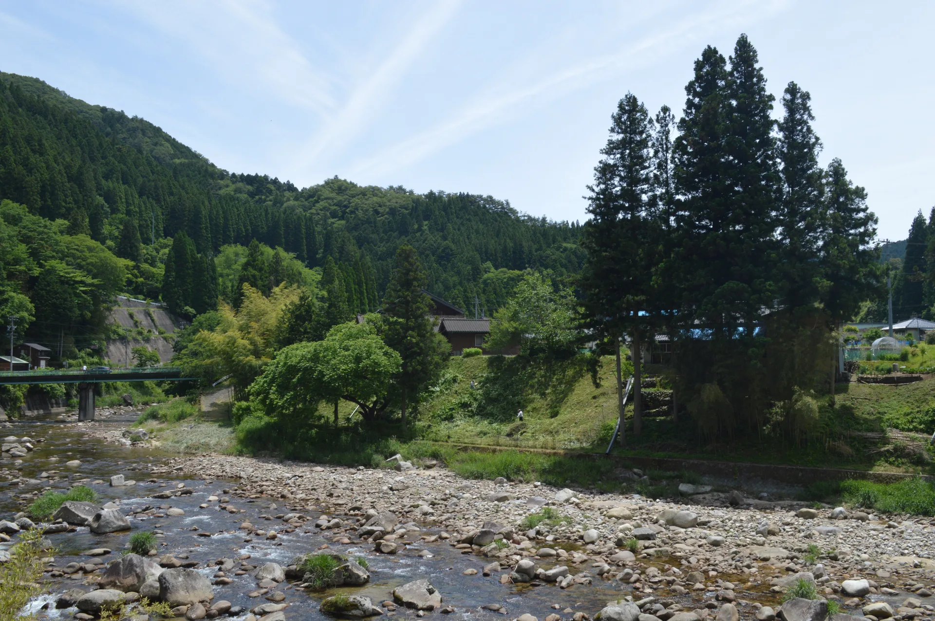 The rocky Neba River flowing below a small bridge, village houses and green forest in Tsukize.